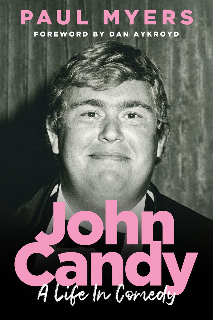 Cover: John Candy: A Life in Comedy, by Paul Myers. Foreword by Dan Aykroyd. A black and white photograph of the comedian and actor John Candy, shown from the chest up, wearing a tuxedo, and looking directly into the camera, with just the hint of a smile. The main title and author’s name appear in large, pink block letters. The sub-title appears in a white, handwritten typeface. The foreword attribution appears in the same block letters as the title, but smaller and in white. 