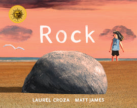  Cover: Rock. Written by Laurel Croza and illustrated by Matt James. The illustrations were created with acrylic paint. A round gray rock sits in the foreground on a sandy beach. The perspective of the illustration makes the rock look large. In the background a young child with  light skin tone stands in the sand. A white seagull is flying in the distance. The blue water can be seen on the horizon. The rock is casting a shadow to the left and the sky is a peachy orange. 