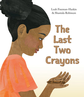  Cover: The Last Two Crayons by Leah Freeman-Haskin and Shantala Robinson. A girl shown in profile smiles as she looks down at two crayons in her hand. Her head and upper body are visible. She has medium skin tone and black hair. She wears a reddish-orange top with short sleeves. The crayons are light brown and dark brown. 