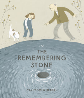  The Remembering Stone by Carey Sookocheff. A girl and an older man with light skin tone, and a white and brown dog, stand on the edge of a body of water, facing us. A stone the man has just thrown skips toward us. The girl has shoulder-length brown hair with a hair bow, and wears blue overalls, a long-sleeved white and grey striped shirt, and tan shoes. The man is bald with grey hair on the side of his head, and wears a brown cardigan over a white collared shirt, grey checked pants, and grey shoes. 