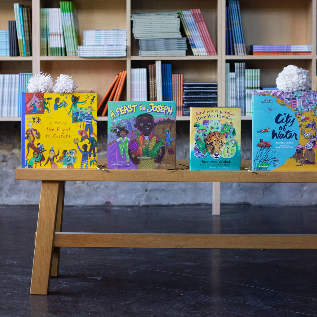  A photo of four books sitting upright on a wooden bench. The books are: Aquí era el paraíso / Here Was Paradise, A Feast for Joseph, I Have the Right to Culture, and City of Water. 